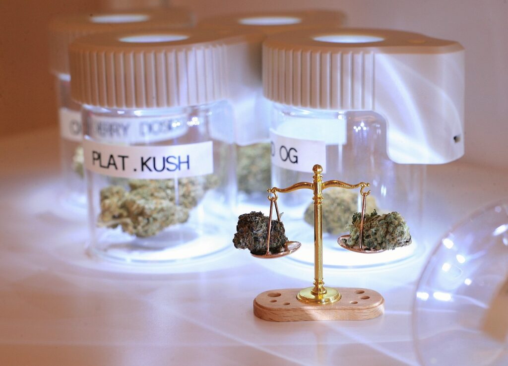 Several jars containing different strains of cannabis, labeled "CHERRY DO," "PLAT. KUSH," and "OG," are arranged in the background. In the foreground, a small golden scale balances THC and CBD buds on each side. The scene is well-lit, emphasizing the display.