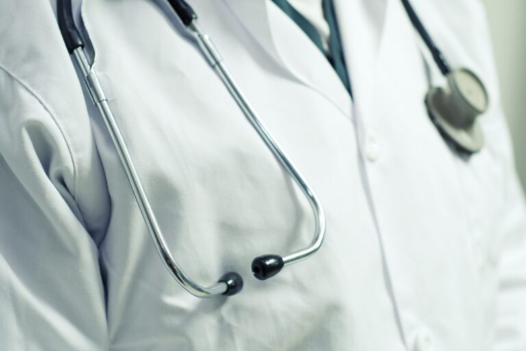 Close-up of a Marijuana Doctor wearing a white lab coat with a stethoscope draped around their neck. The stethoscope's earpieces and part of the chest piece are visible, while the background is blurred and in neutral tones.